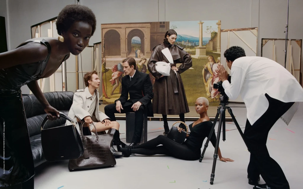 Fashion editorial featuring six diverse models in a studio setting with designer handbags and a photographer capturing the scene against a painted backdrop.