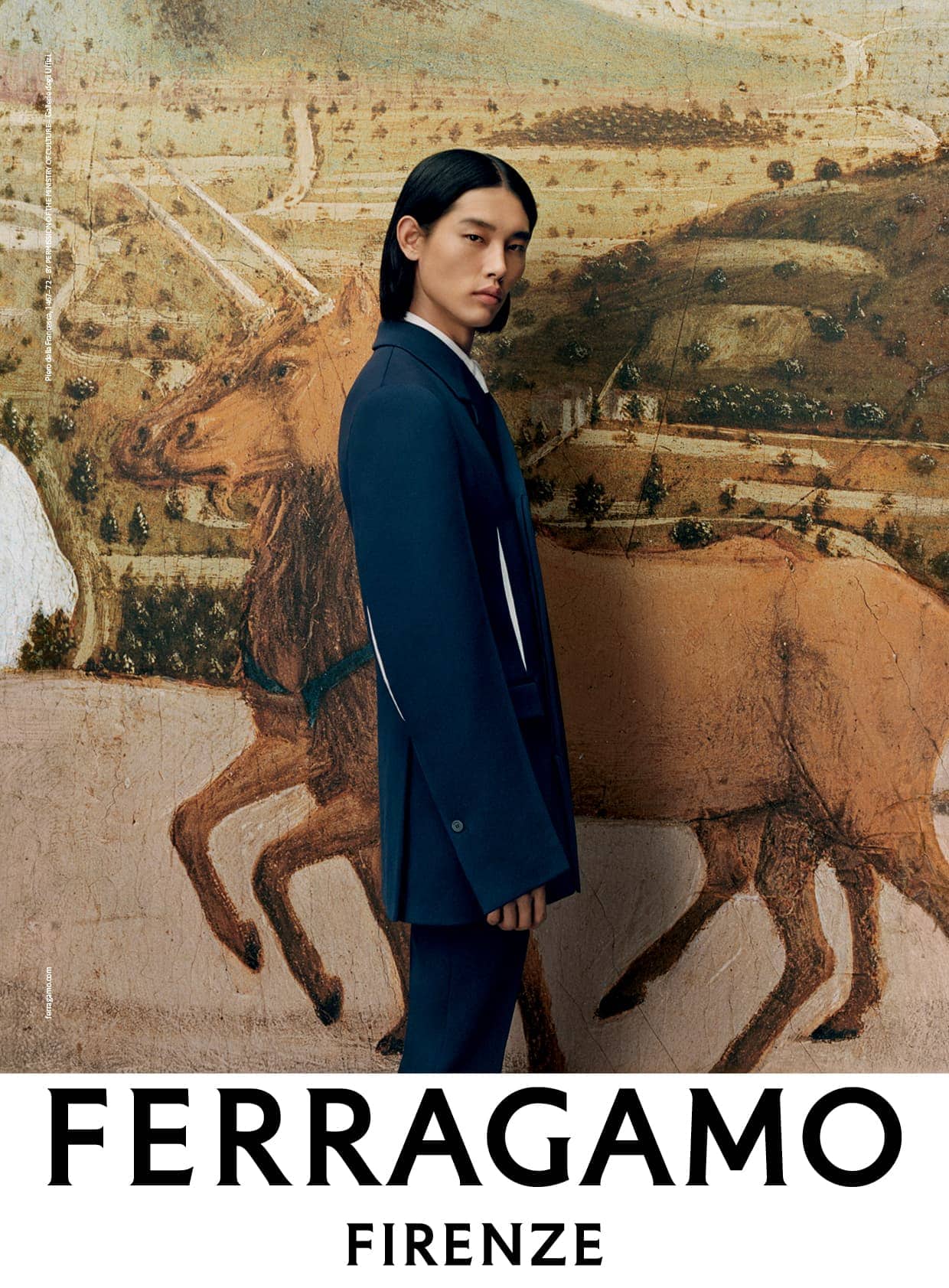 A young Asian man poses in a dark blue suit against a backdrop of a light brown and tan Renaissance painting of mounted figures.