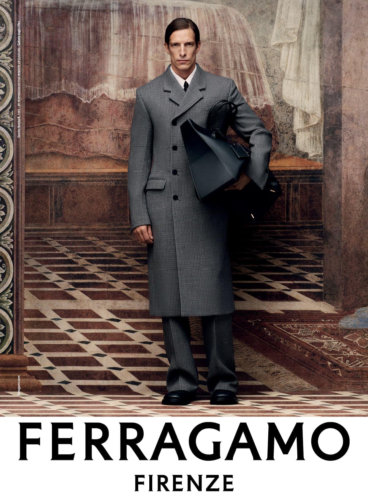 Fashion model wears grey coat and trousers, carrying a black bag in an indoor setting with a painted backdrop and tiled floor for an advertisement.