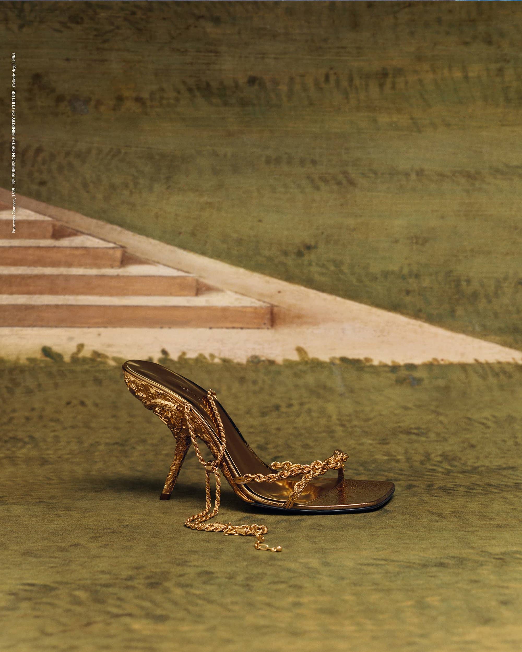 Elegant golden sandal with chain detail showcased against backdrop of stairs and textured green surface in a fashion still life.