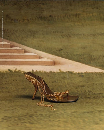 Elegant golden sandal with chain detail showcased against backdrop of stairs and textured green surface in a fashion still life.