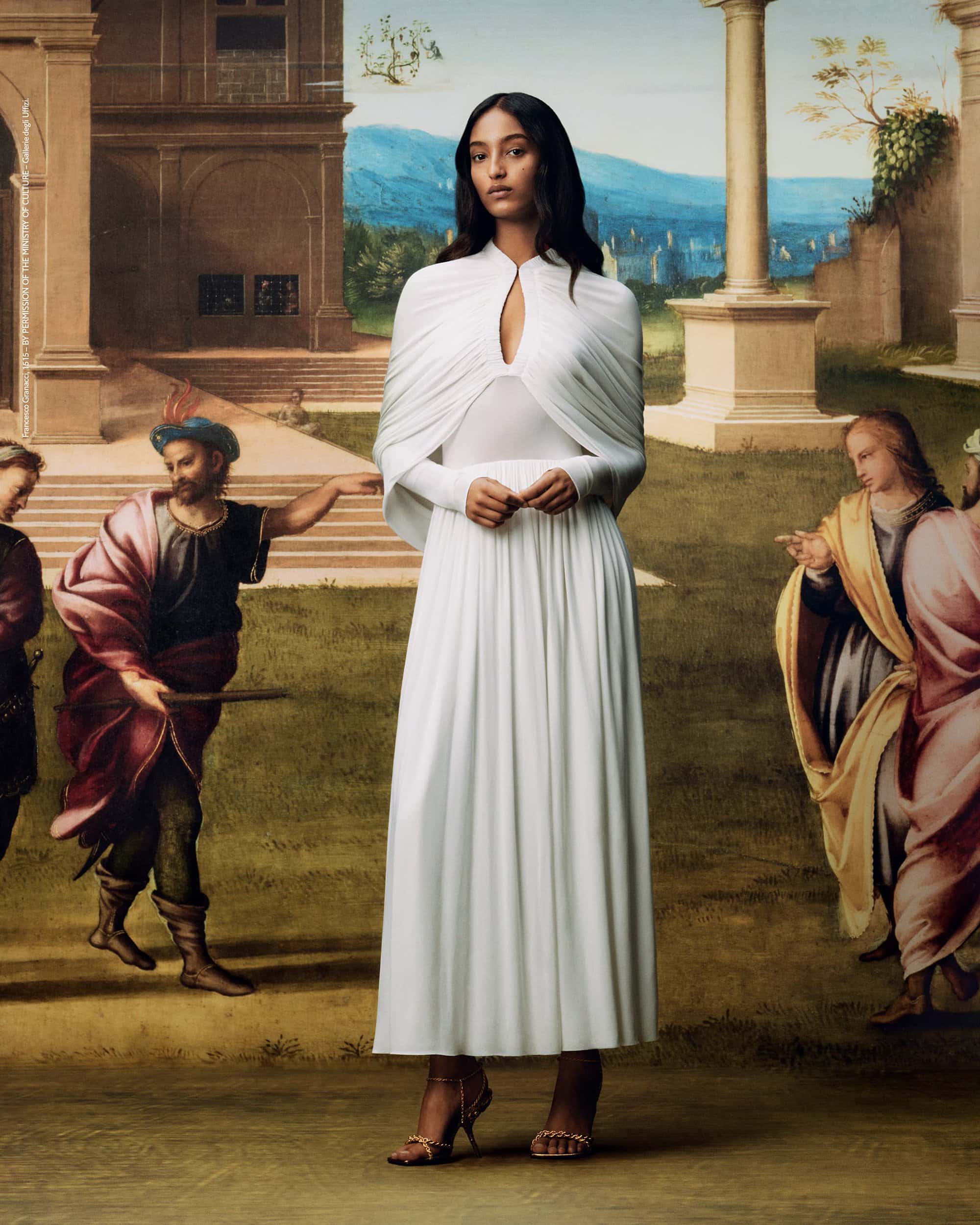 Elegant woman in a white dress and cape stands in front of classical painting backdrop. Neutral tones and soft lighting.