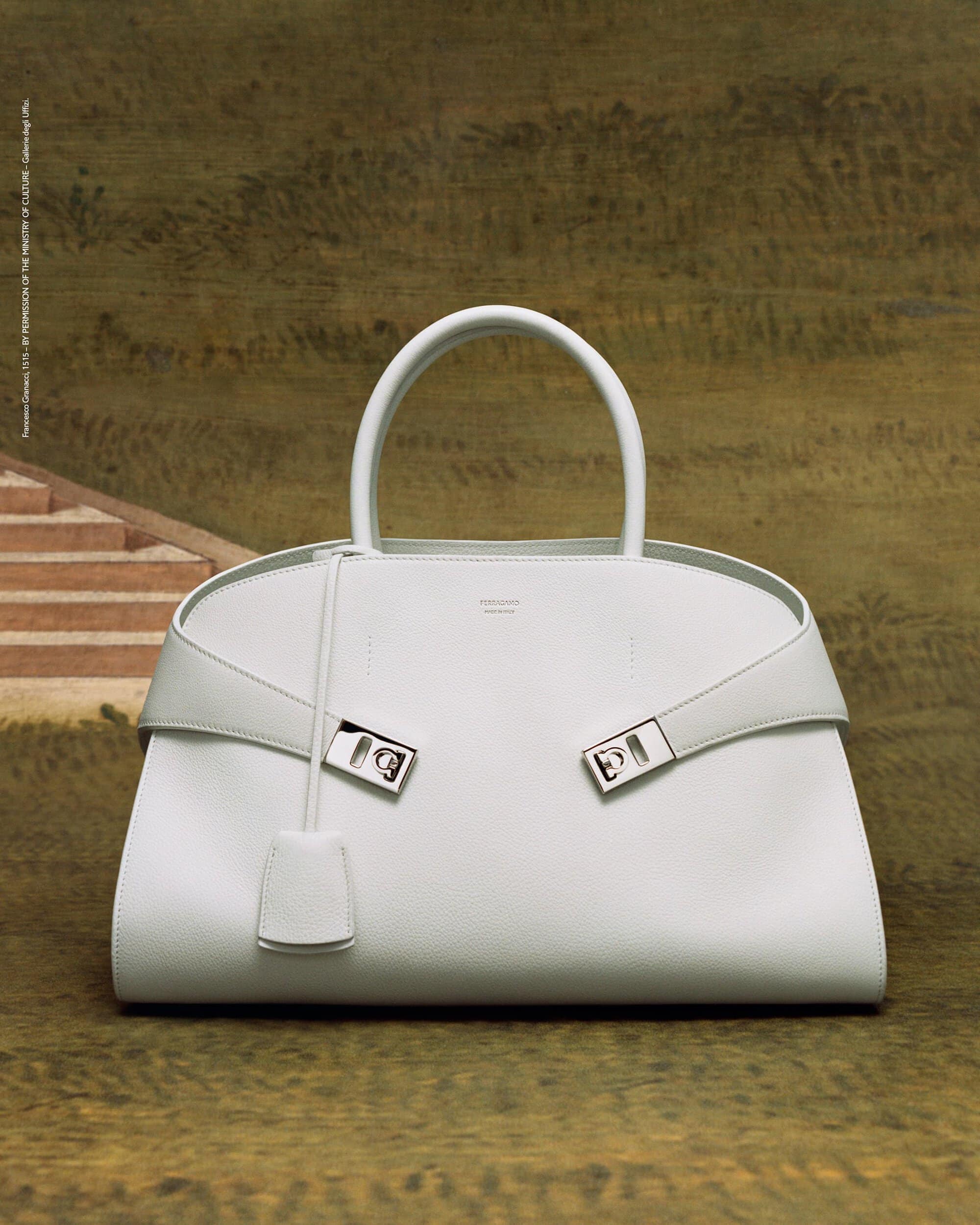 Elegant white leather handbag with silver hardware and top handle against a muted olive and beige scenic backdrop. Fashion accessory.
