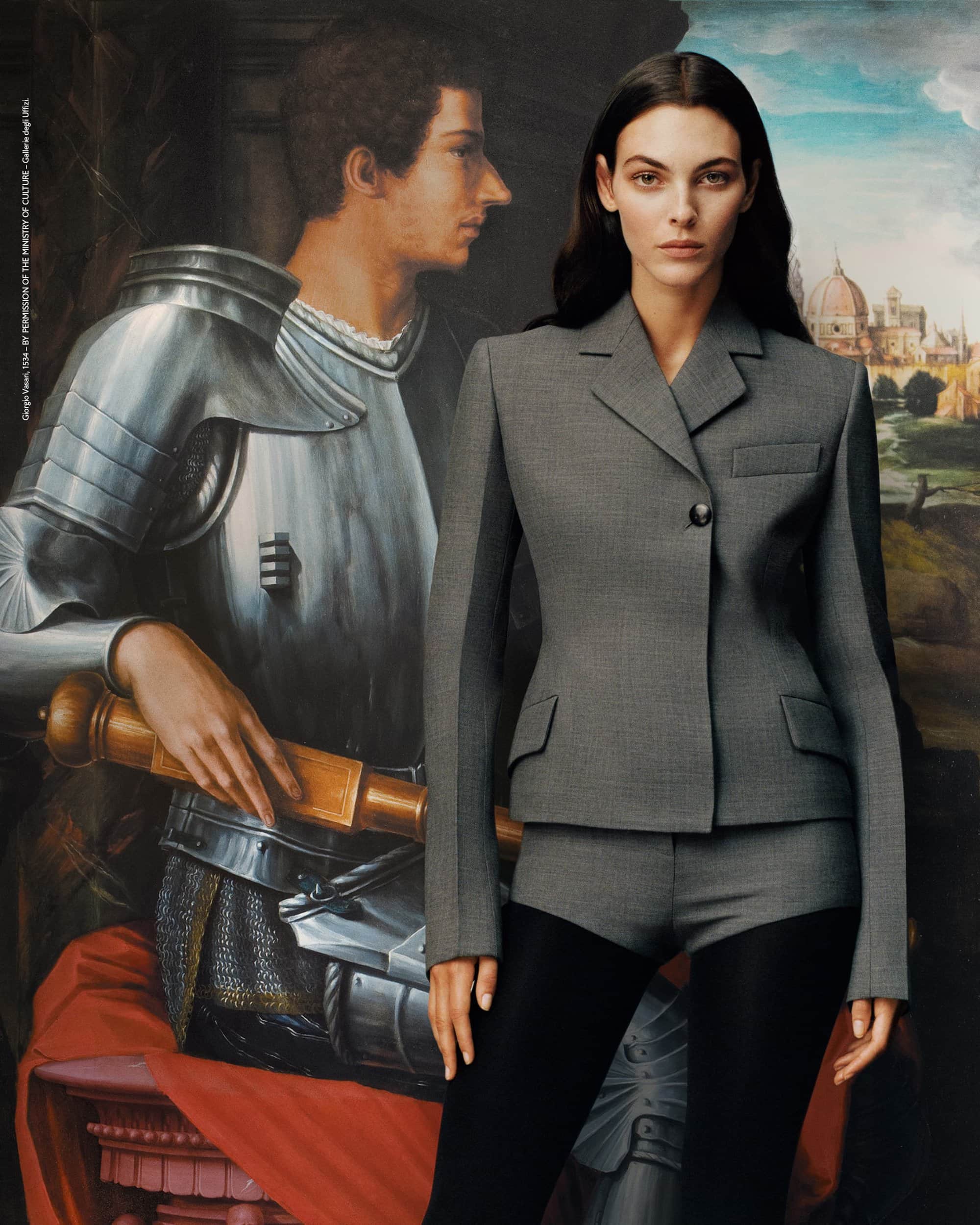 Woman poses in a grey tailored jacket and shorts with black tights, set against a backdrop of a Renaissance-style painting.