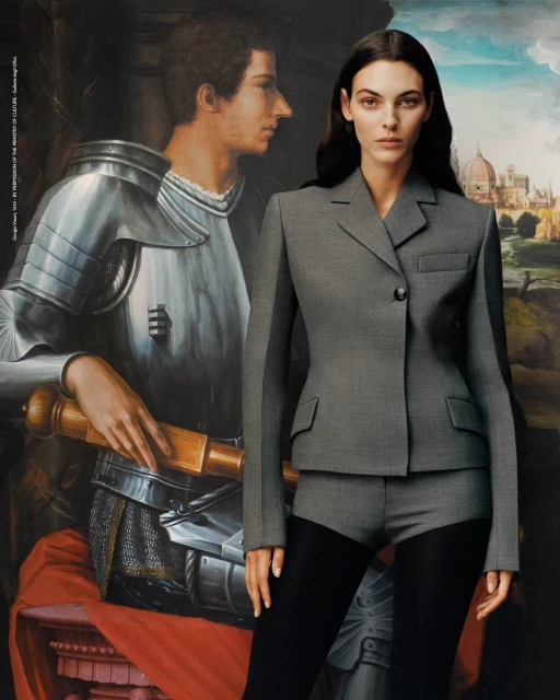 Woman poses in a grey tailored jacket and shorts with black tights, set against a backdrop of a Renaissance-style painting.