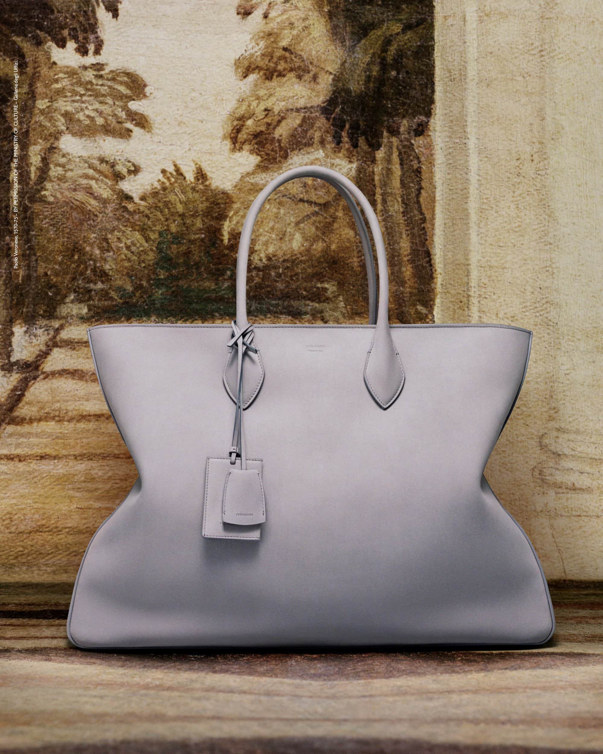 Studio shot of a light grey leather tote bag with handles and tag, set against a blurred vintage painting backdrop.