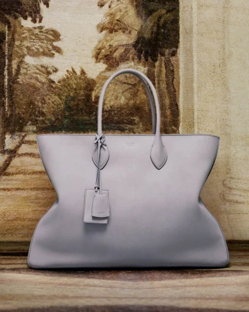 Studio shot of a light grey leather tote bag with handles and tag, set against a blurred vintage painting backdrop.