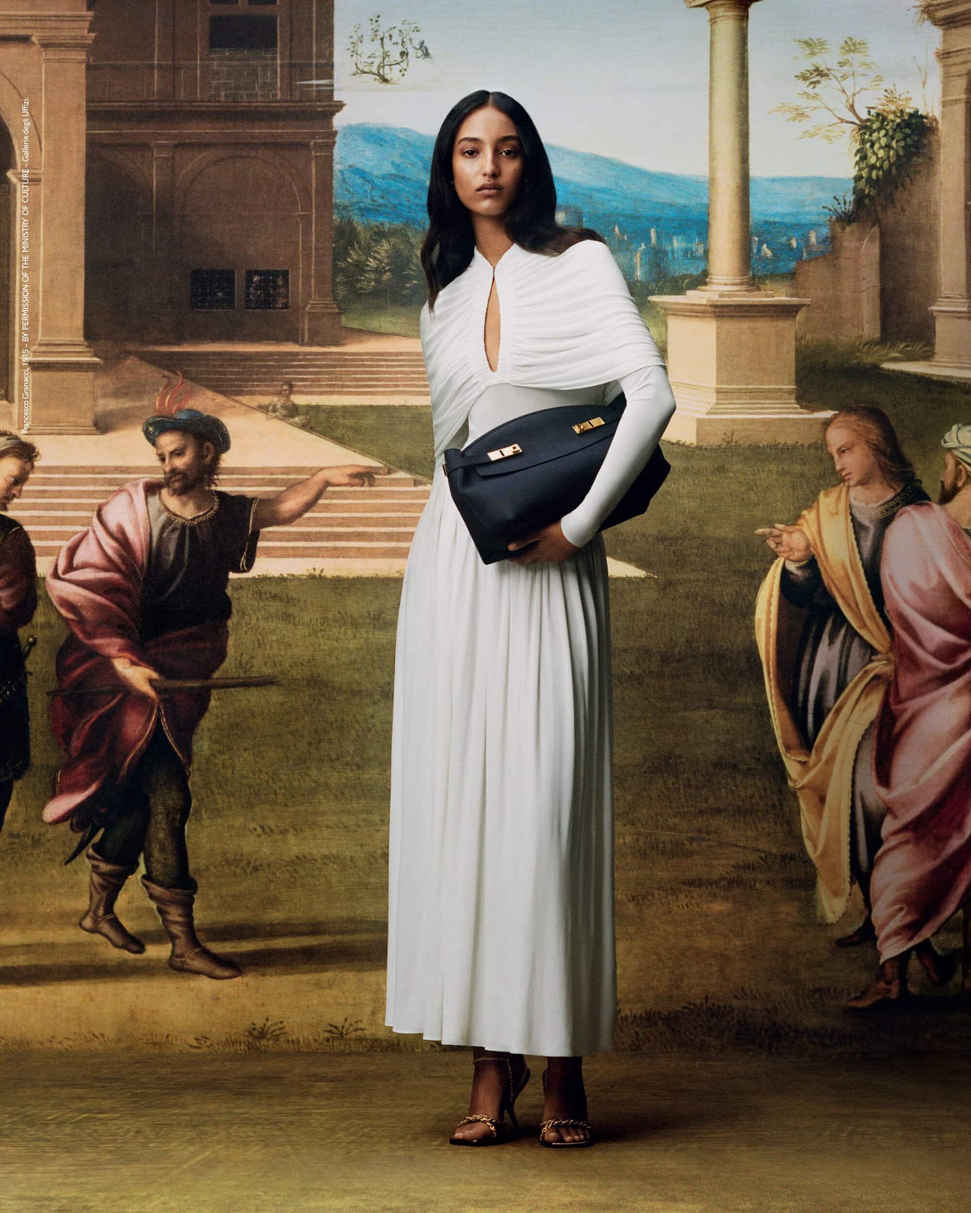 Elegant woman in a white dress holds a black handbag against a backdrop of a Renaissance-era mural featuring classic figures.
