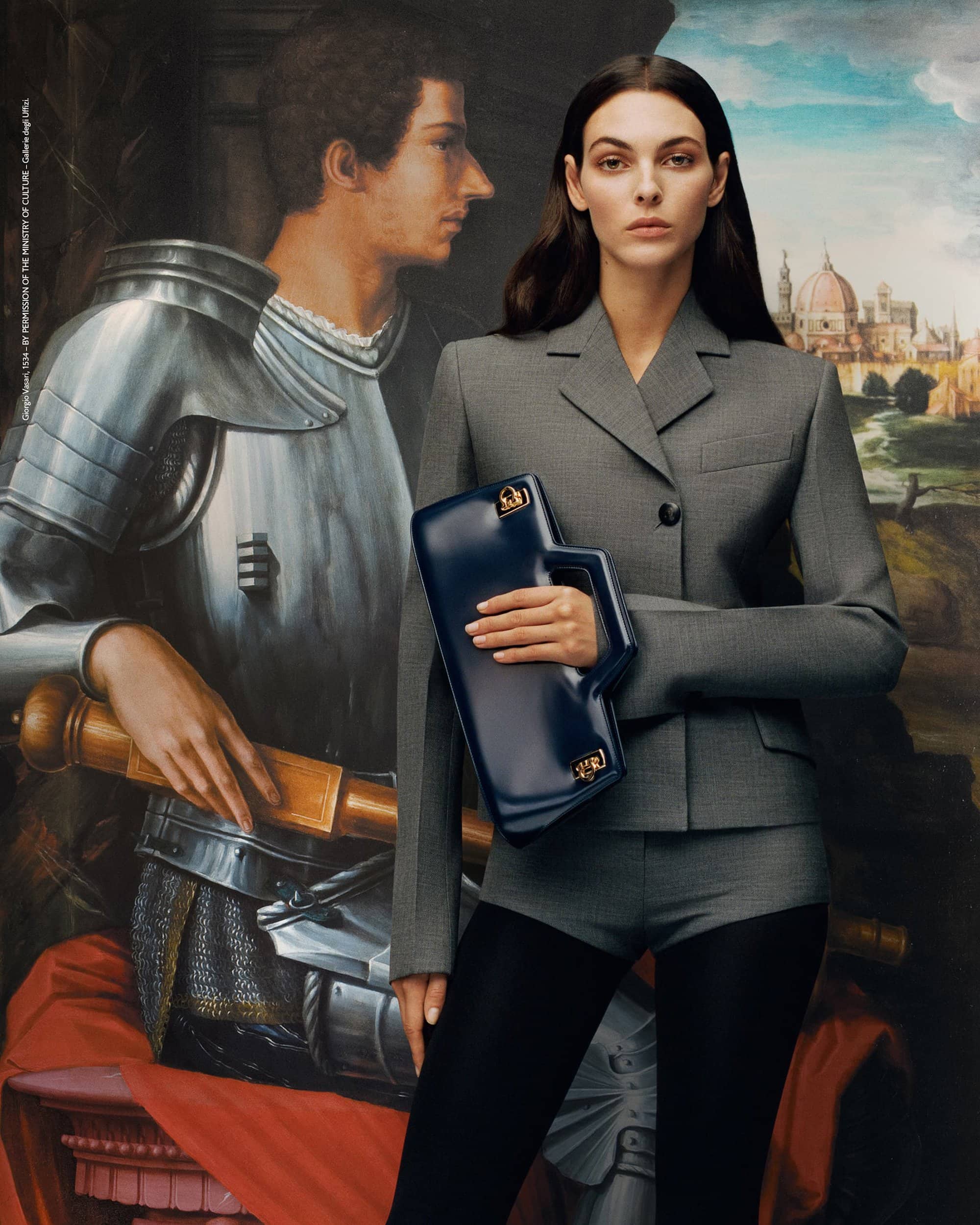 Woman in a gray suit poses with a structured blue handbag in front of an oil painting of a knight and Florence cityscape.