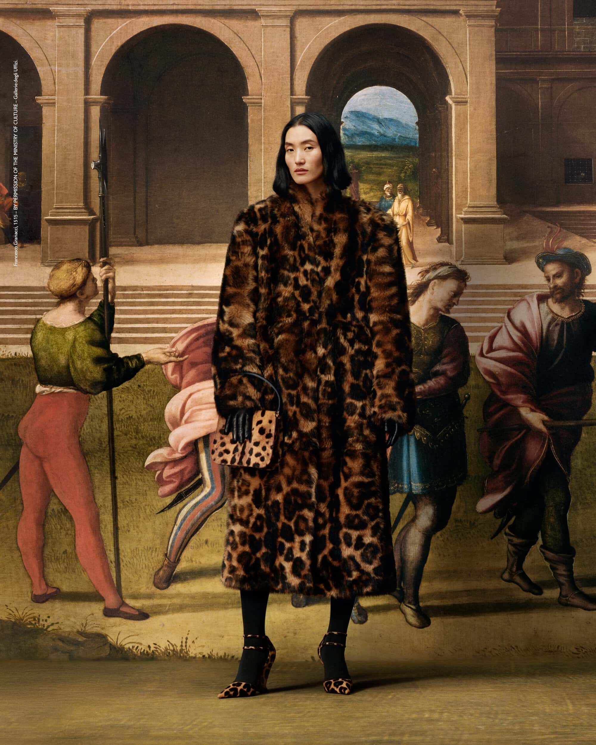 A woman in a full-length leopard print coat poses in front of a Renaissance-style mural, accented by matching leopard print bag and shoes.
