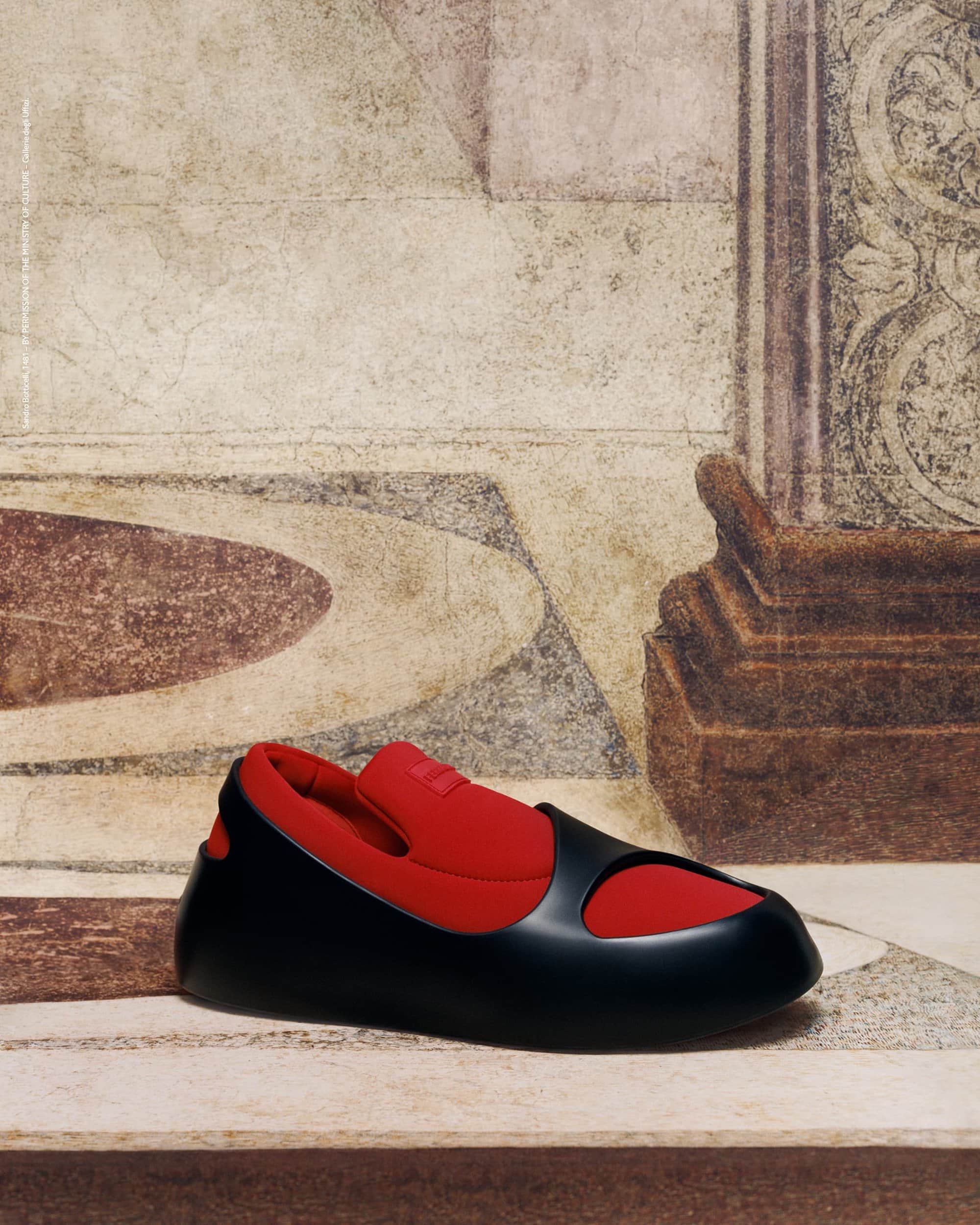 Black and red slip-on shoe against a mottled cream and brown background. The shoe has a rounded toe and a thick sole.