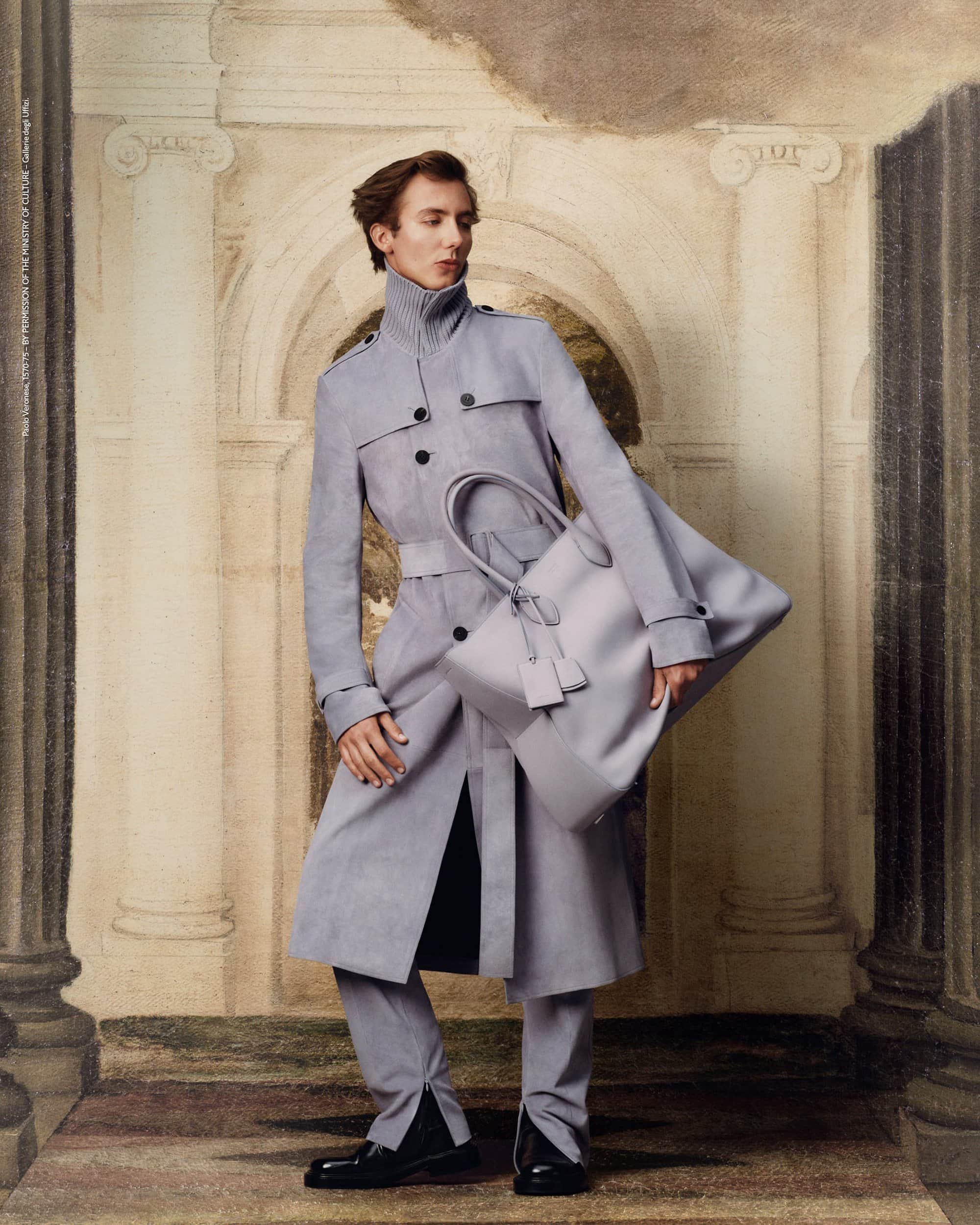 A slender man in a gray trench coat holds a matching handbag against a painted architectural backdrop in a fashion editorial.