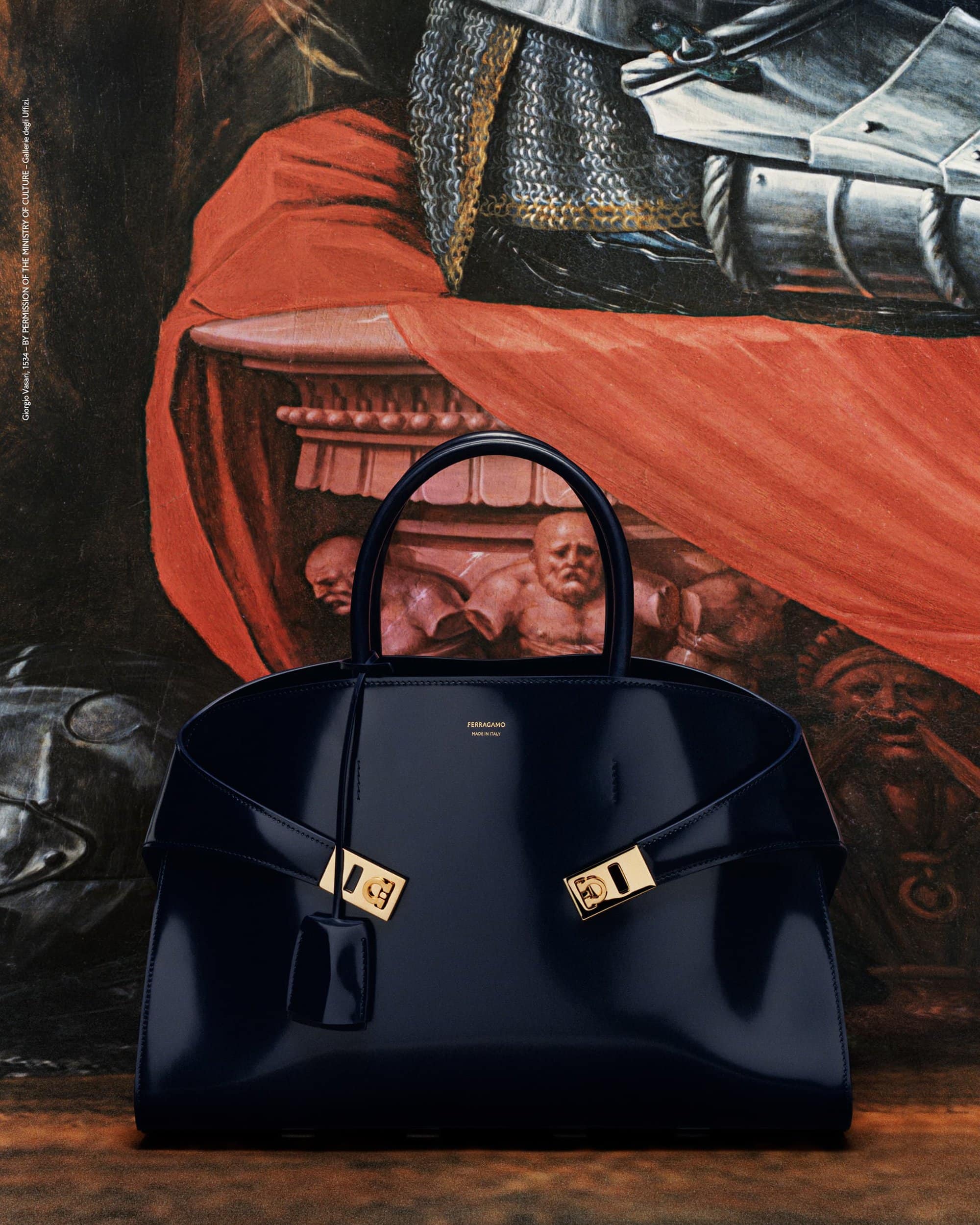 Elegant navy blue handbag with gold hardware in front of an artistic backdrop featuring figures and drapery in a rich, moody palette.