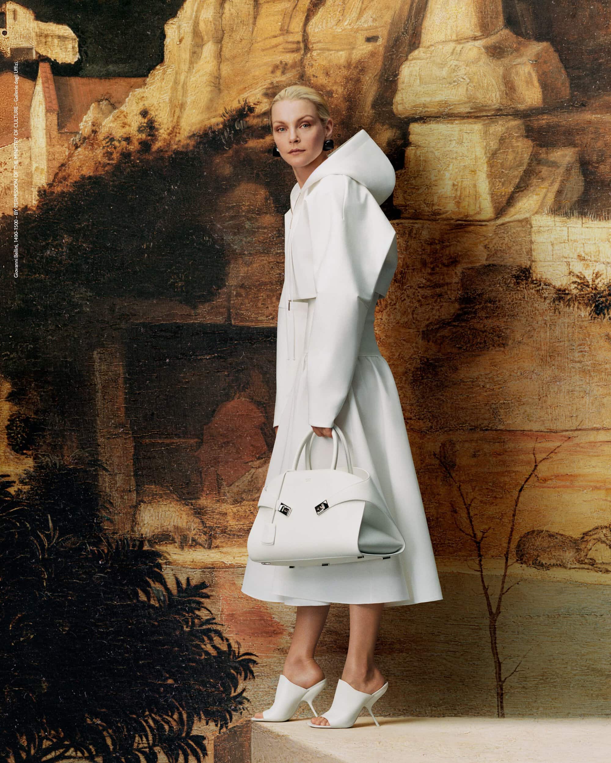 Fashion editorial shot of a blonde woman in a white hooded dress, holding a structured white handbag against a Renaissance-style backdrop.