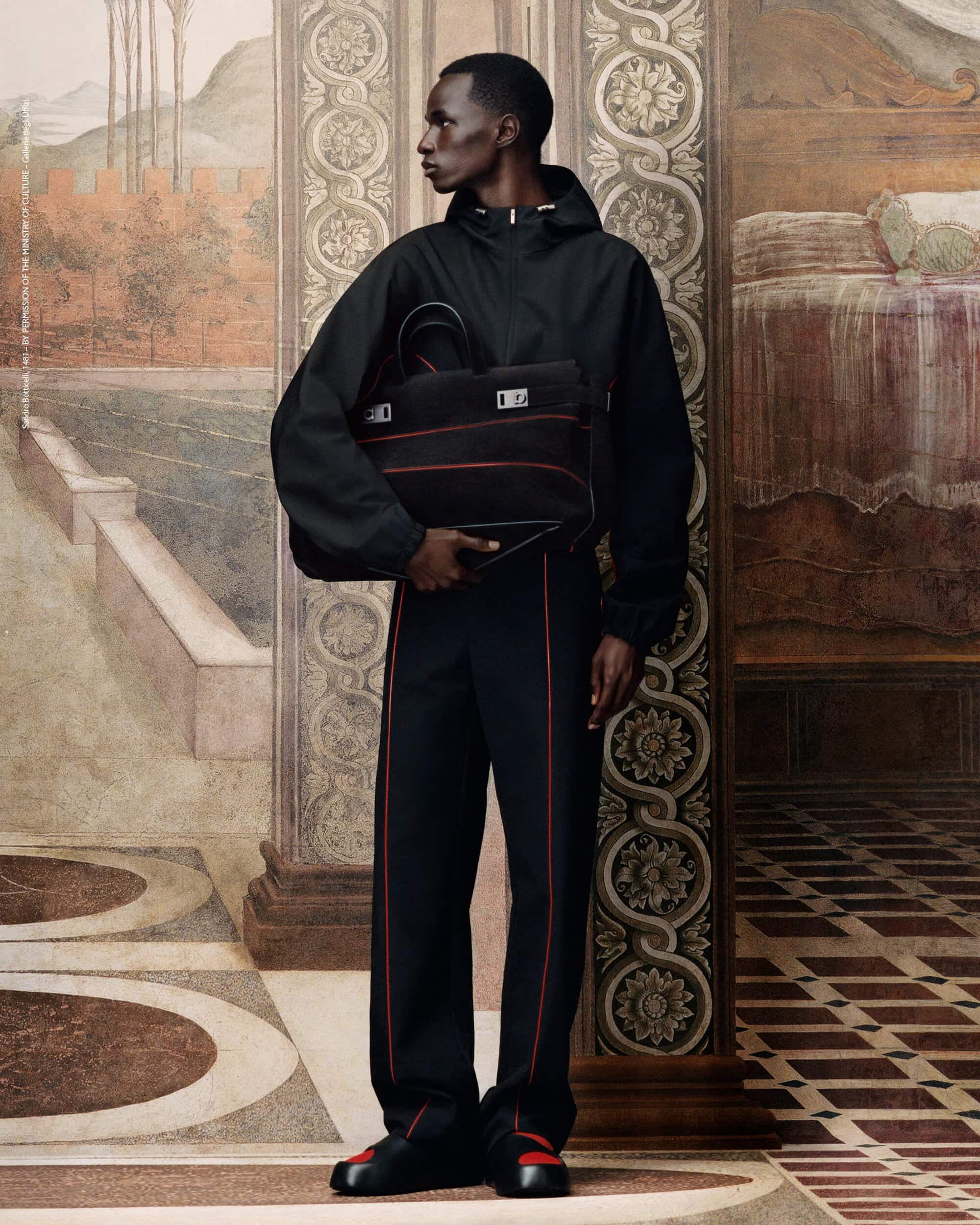 Fashionable Black man in anorak and track pants holds a black tote bag against an abstract studio backdrop with muted earth tones.