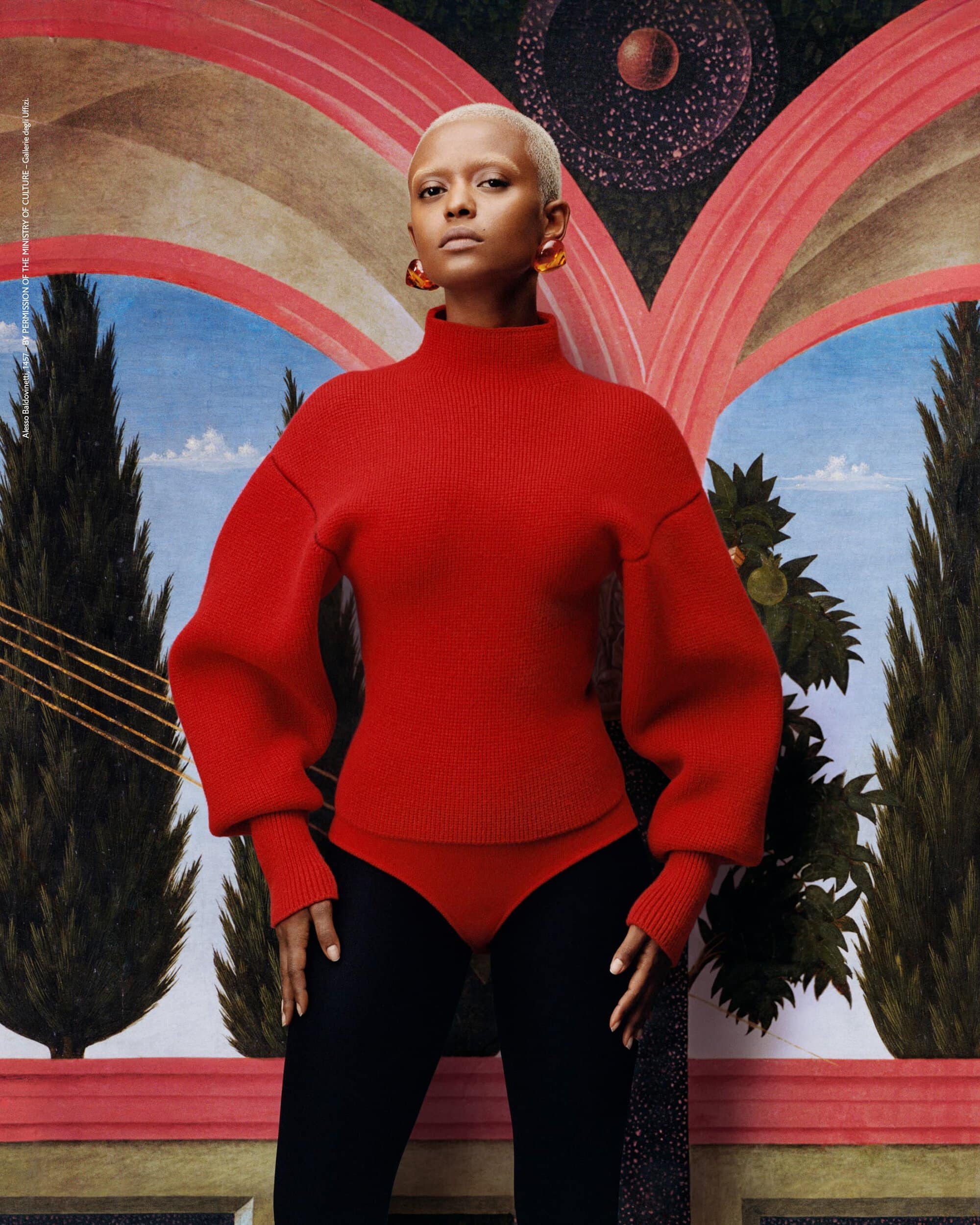 Woman with shaved head in red sweater and black leggings posing in front of an artistic backdrop, fashion editorial style image.