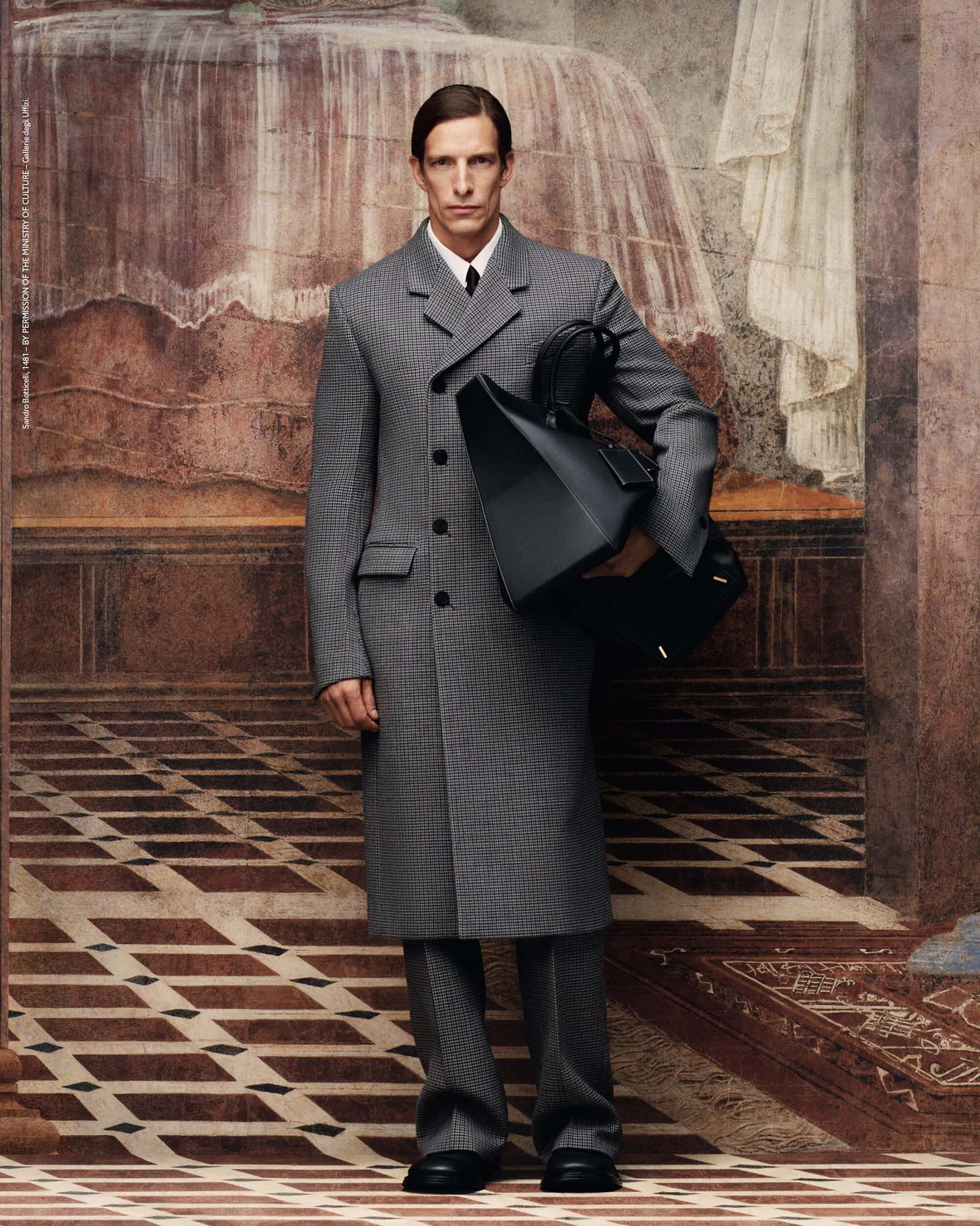 Male model in a grey striped suit and overcoat, holding a black structured bag against a backdrop with mural and tiled floor.