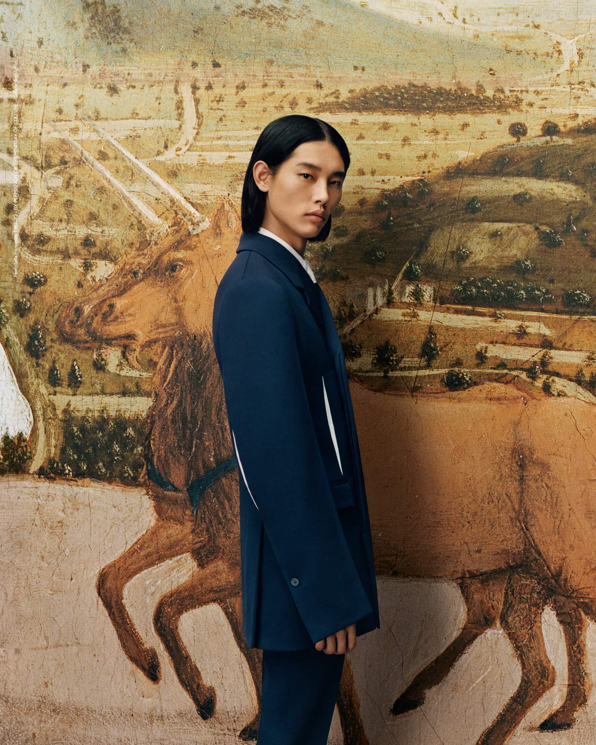 An adult male model with long black hair wearing a blue suit poses against an artistic backdrop with an image of a brown horse.