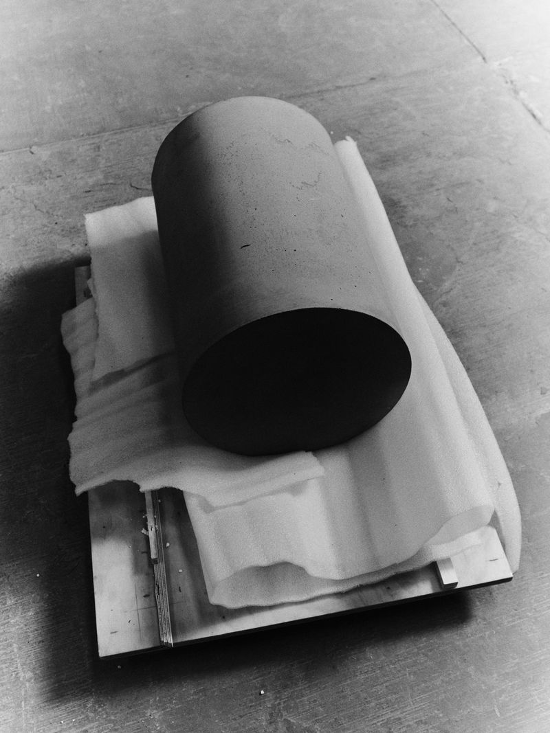 A cylindrical object rests on a cushioned surface atop a wooden board in a stark black and white still life product photograph.