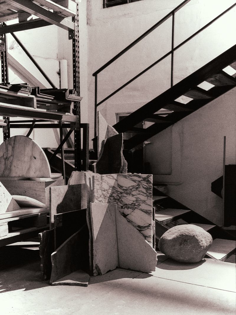 Black and white photograph of assorted marble and stone pieces piled on a concrete floor, with stairs in the background.