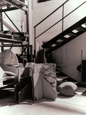 Black and white photograph of assorted marble and stone pieces piled on a concrete floor, with stairs in the background.