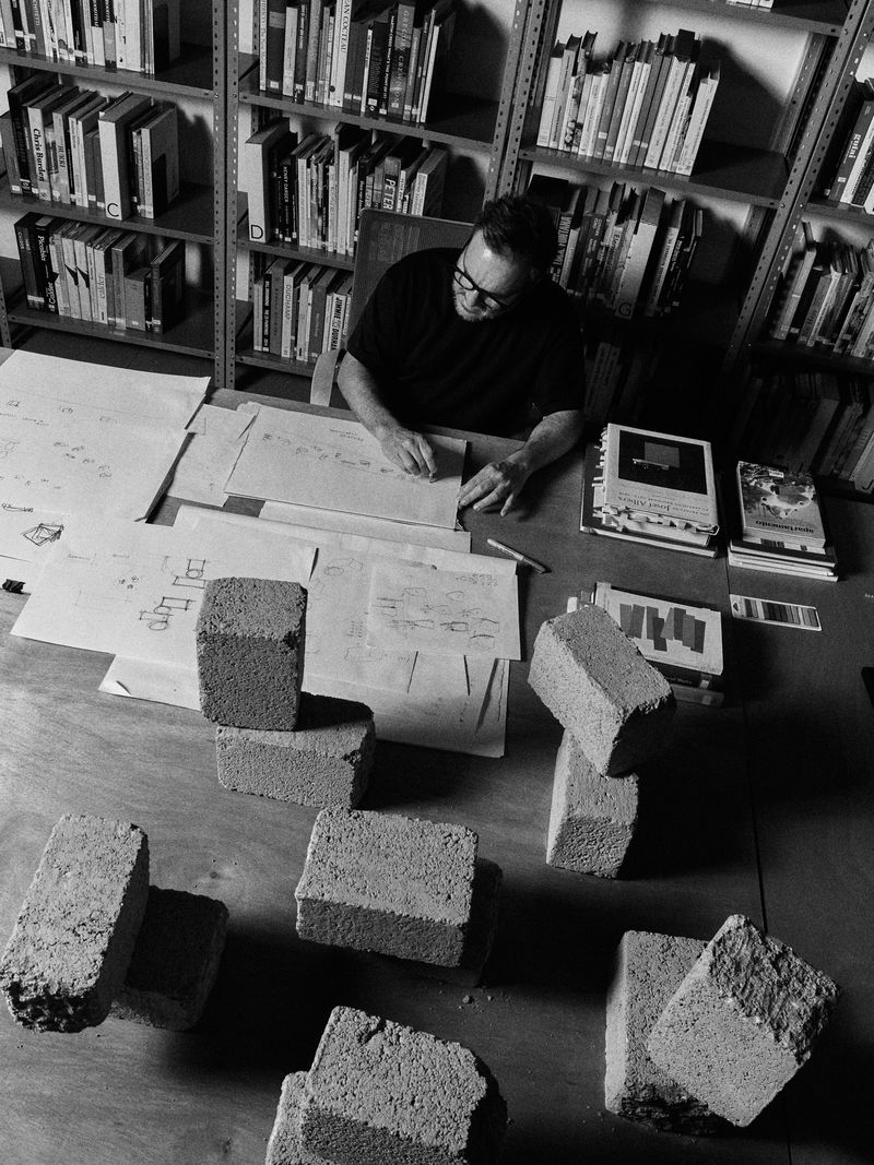 Black-and-white image of a man with glasses at a desk, drawing on papers surrounded by bricks, with bookshelves in the background.