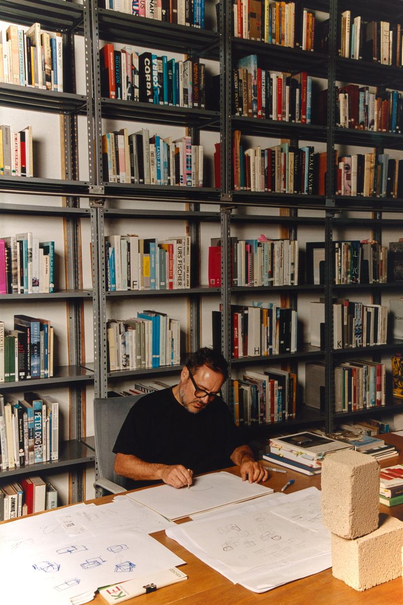 Man drawing architectural sketches at a desk surrounded by books and cinder blocks in a bright indoor office or studio space.