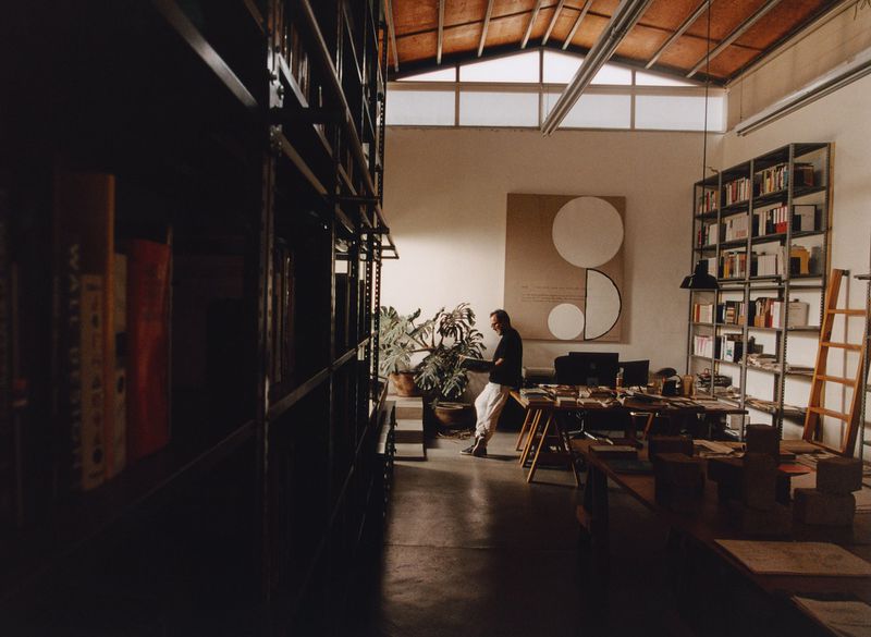 Well-lit modern office space with a man leaning against a table, bookshelves, indoor plants, and a geometric art piece on the wall.