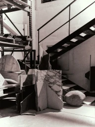 Black and white photograph showing stone slabs, metal shelving, and a dark metal staircase in an industrial workshop interior