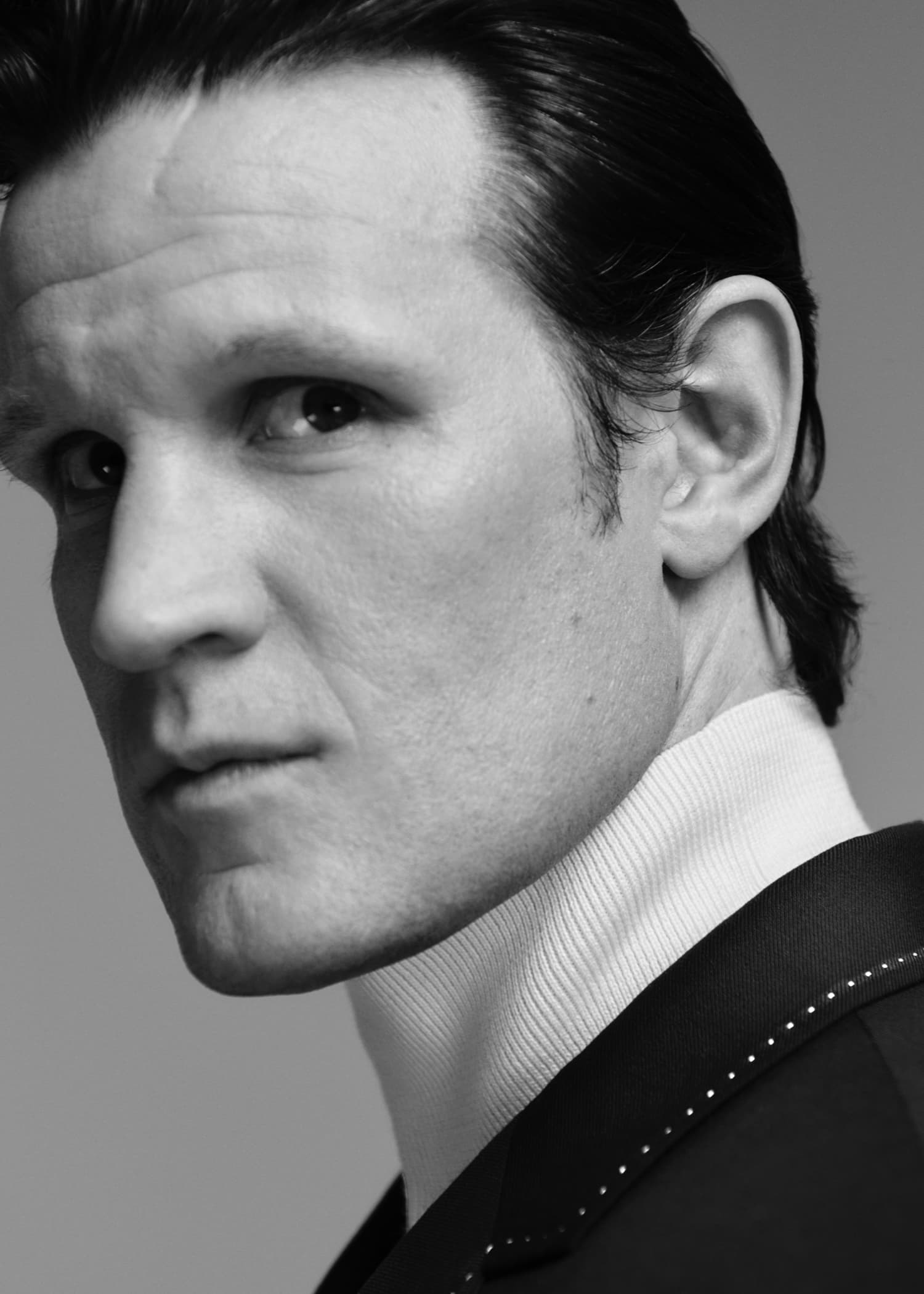Black and white portrait of a male actor with slicked-back hair, wearing a jacket over a high-necked top, in a studio setting.