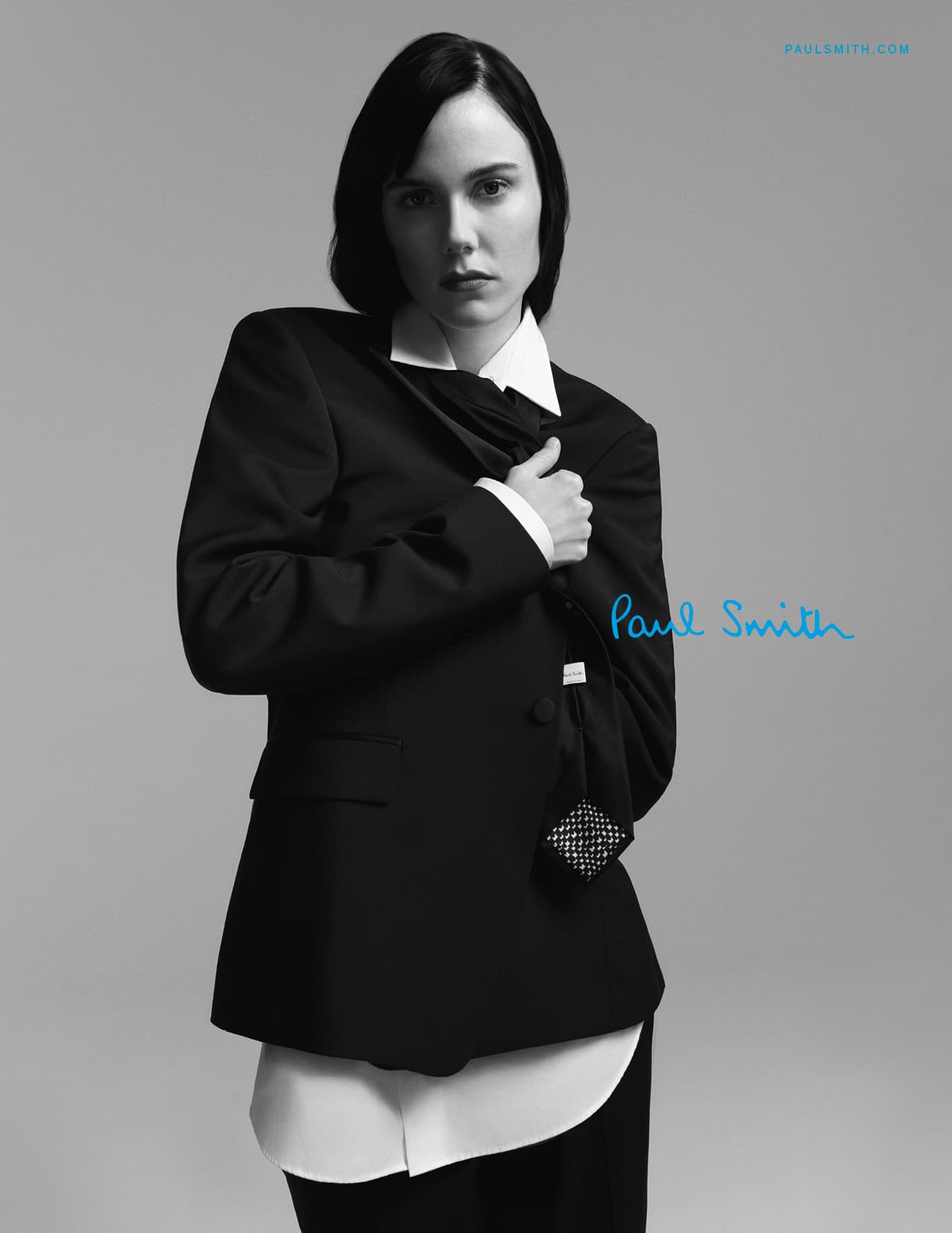 An adult female in black suit and white shirt poses indoors, against a grey studio backdrop. She looks directly at the camera.