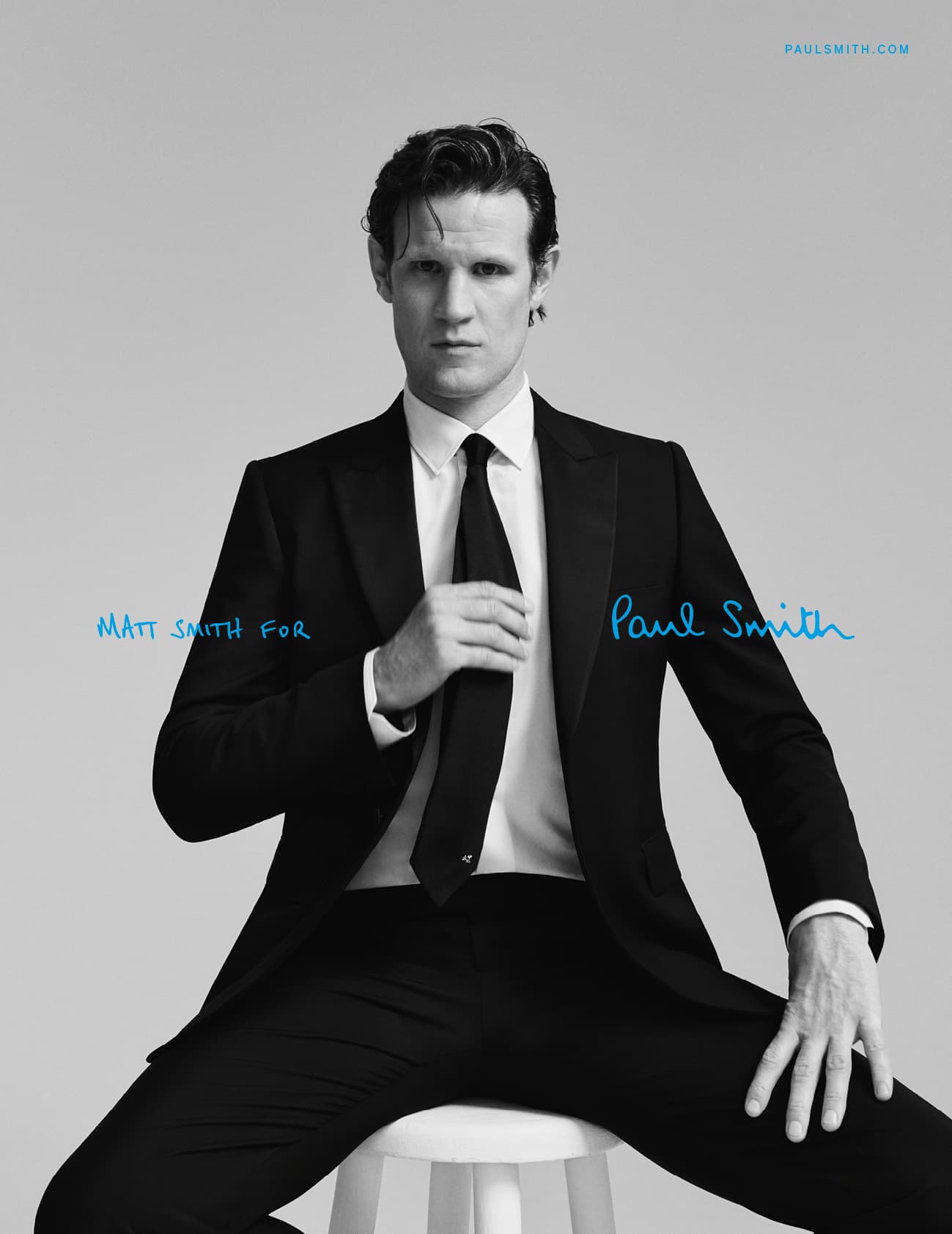 Matt Smith for Paul Smith campaign: Man in black suit sits on stool in studio with blue brand typography in a fashion editorial.