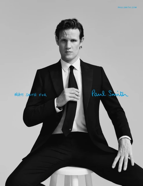 Matt Smith for Paul Smith campaign: Man in black suit sits on stool in studio with blue brand typography in a fashion editorial.