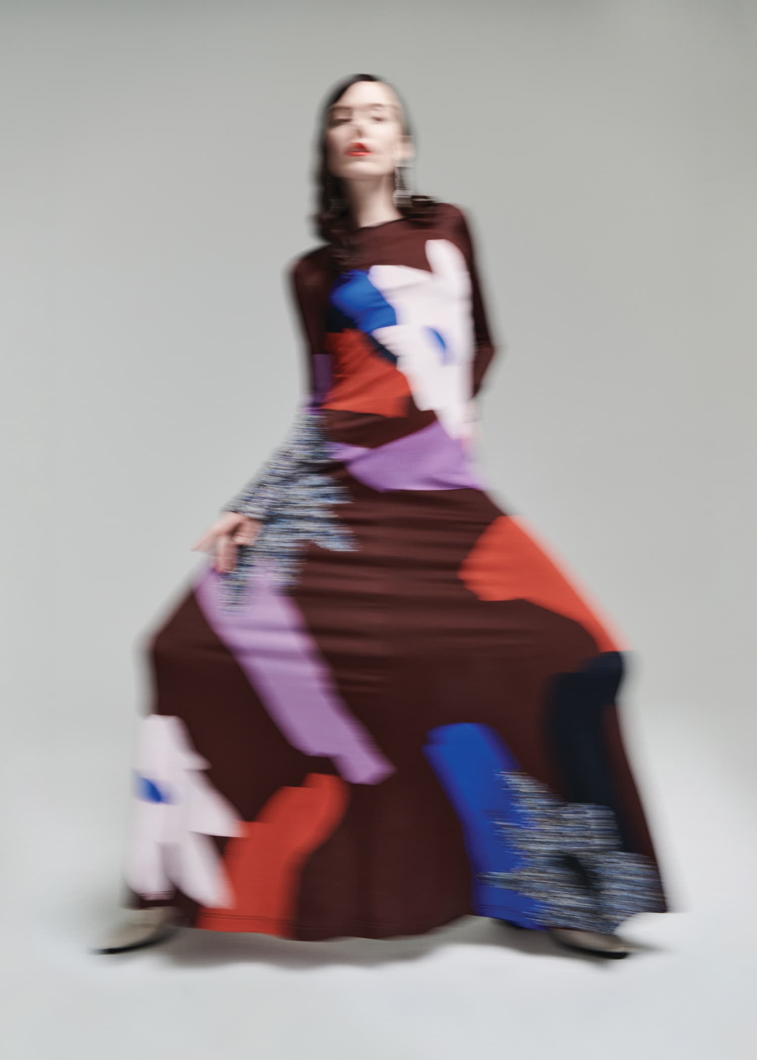 A blurry image of a stylish woman wearing a brown, long-sleeved maxi dress with colorful geometric patterns against a neutral backdrop.
