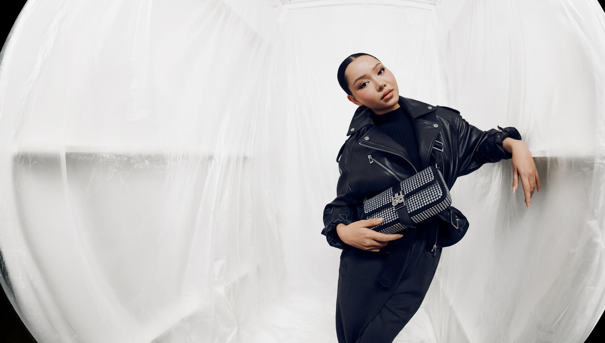 A young woman in a black leather jacket and black pants is posing with a black and silver purse in front of a white, draped backdrop.