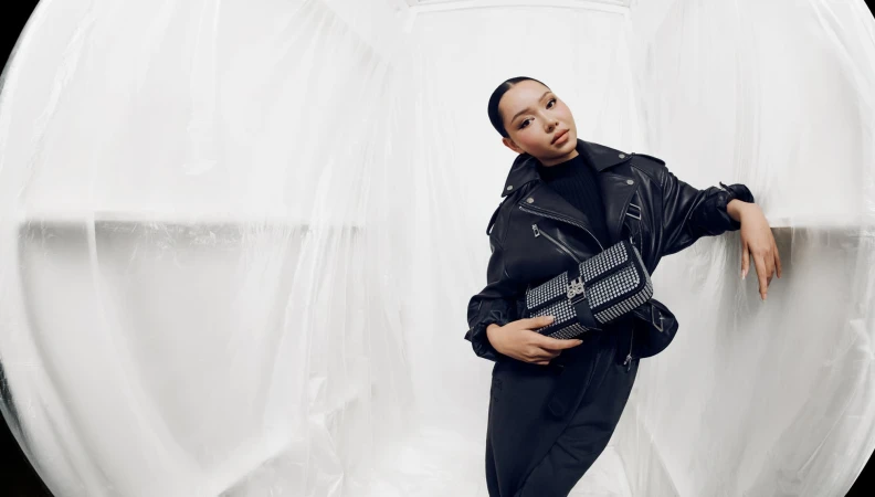 A young woman in a black leather jacket and black pants is posing with a black and silver purse in front of a white, draped backdrop.