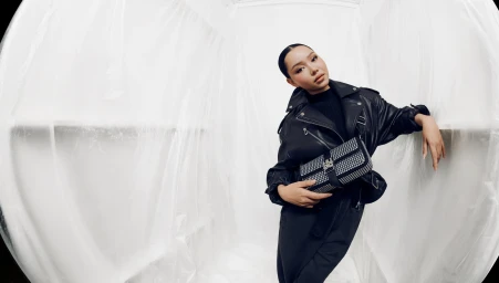 Bella Poarch wearing a black leather jacket and holding an embellished bag in a white plastic draped studio setting.