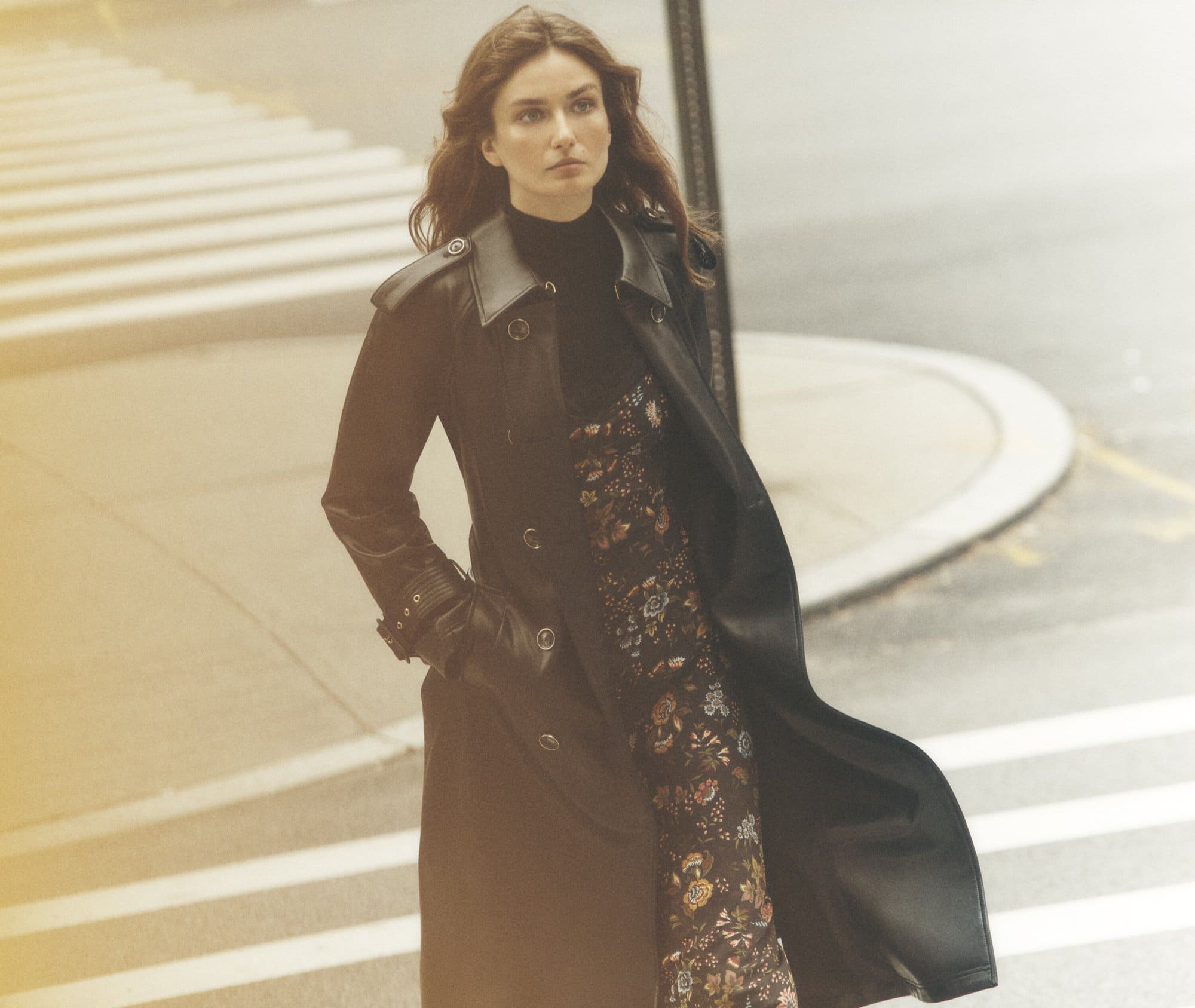 Woman in stylish black coat and floral dress crossing the street with a modern urban background on a sunny day in a city.