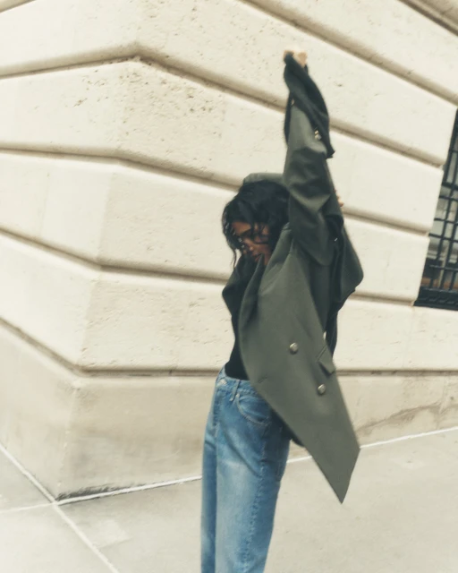 An adult female stands outdoors in an olive jacket and jeans, arms raised against a neutral building backdrop on an overcast day.