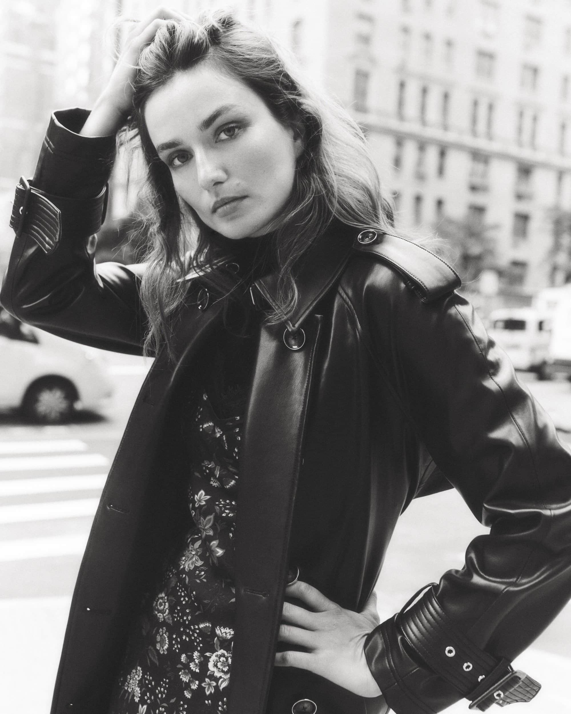 A fashionable woman in a black coat poses in a city street. The photo features a black-and-white aesthetic, enhancing the urban setting.
