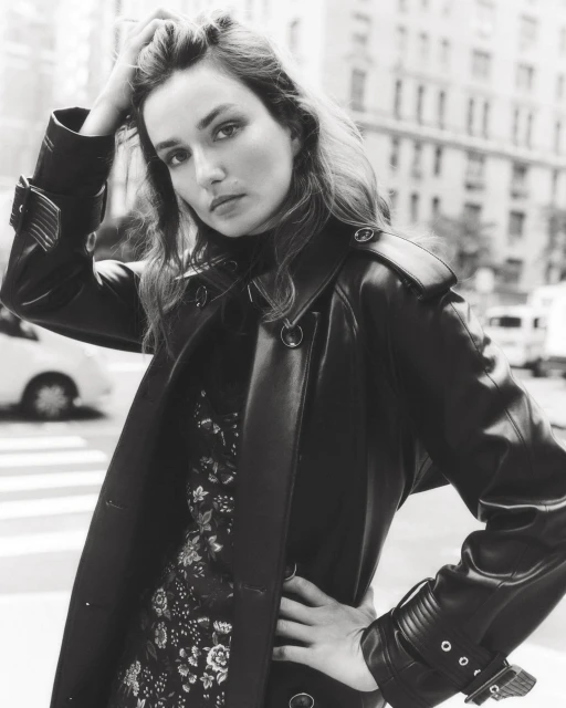 A fashionable woman in a black coat poses in a city street. The photo features a black-and-white aesthetic, enhancing the urban setting.