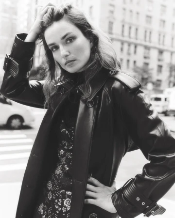 A fashionable woman in a black coat poses in a city street. The photo features a black-and-white aesthetic, enhancing the urban setting.