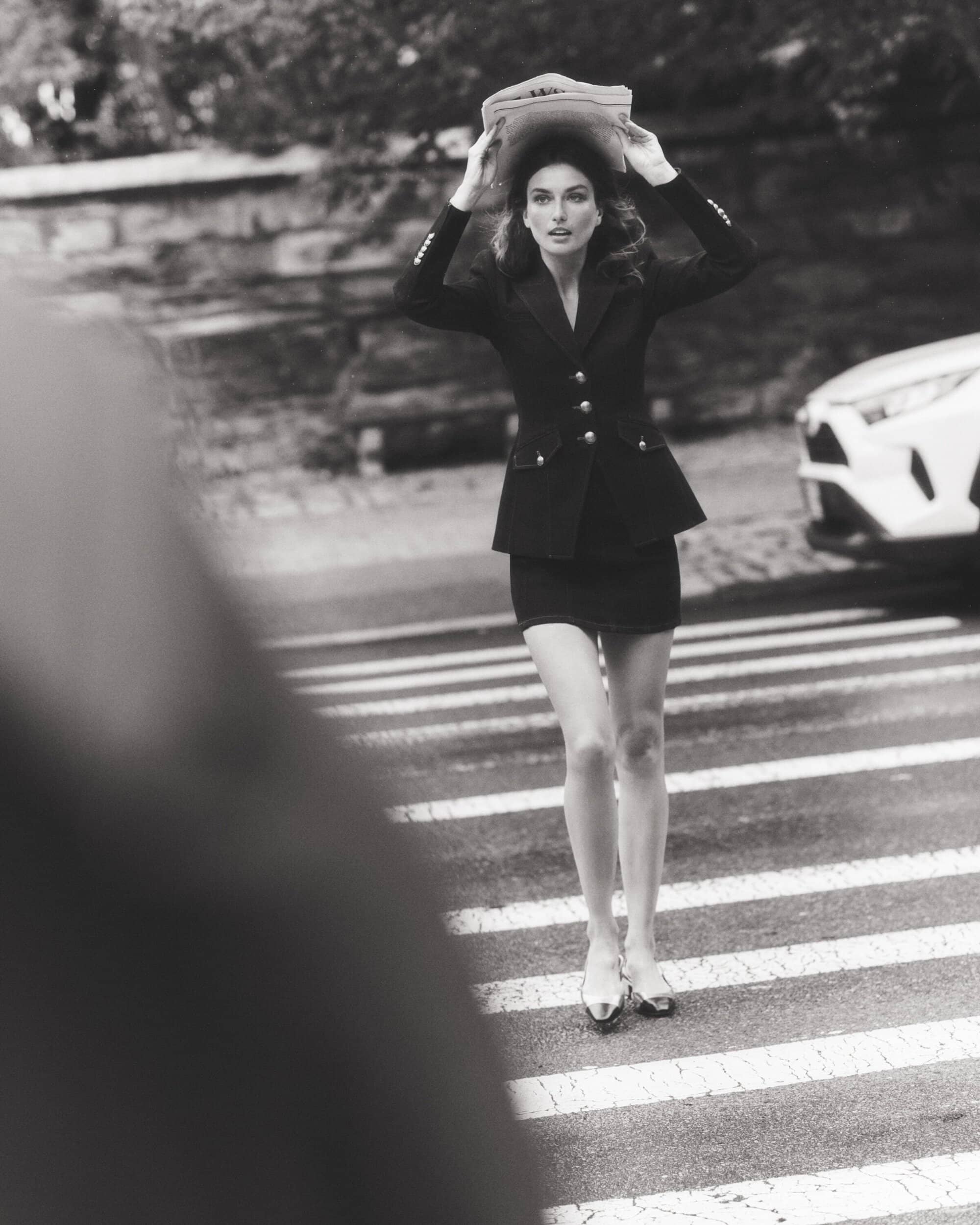 Elegant woman in black skirt suit crossing street with newspaper over head. Black and white fashion photography in urban setting.