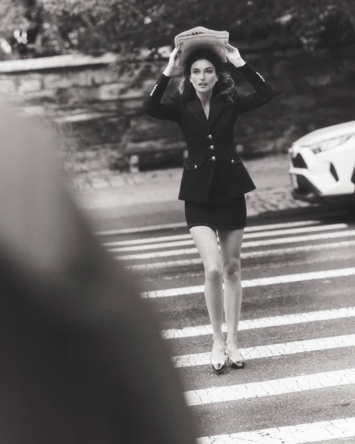 Elegant woman in black skirt suit crossing street with newspaper over head. Black and white fashion photography in urban setting.