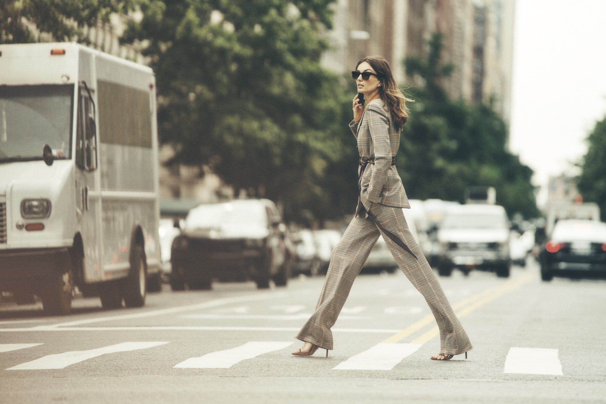 A woman in a plaid suit walks across a crosswalk while talking on her phone, with city buildings and traffic in the background.