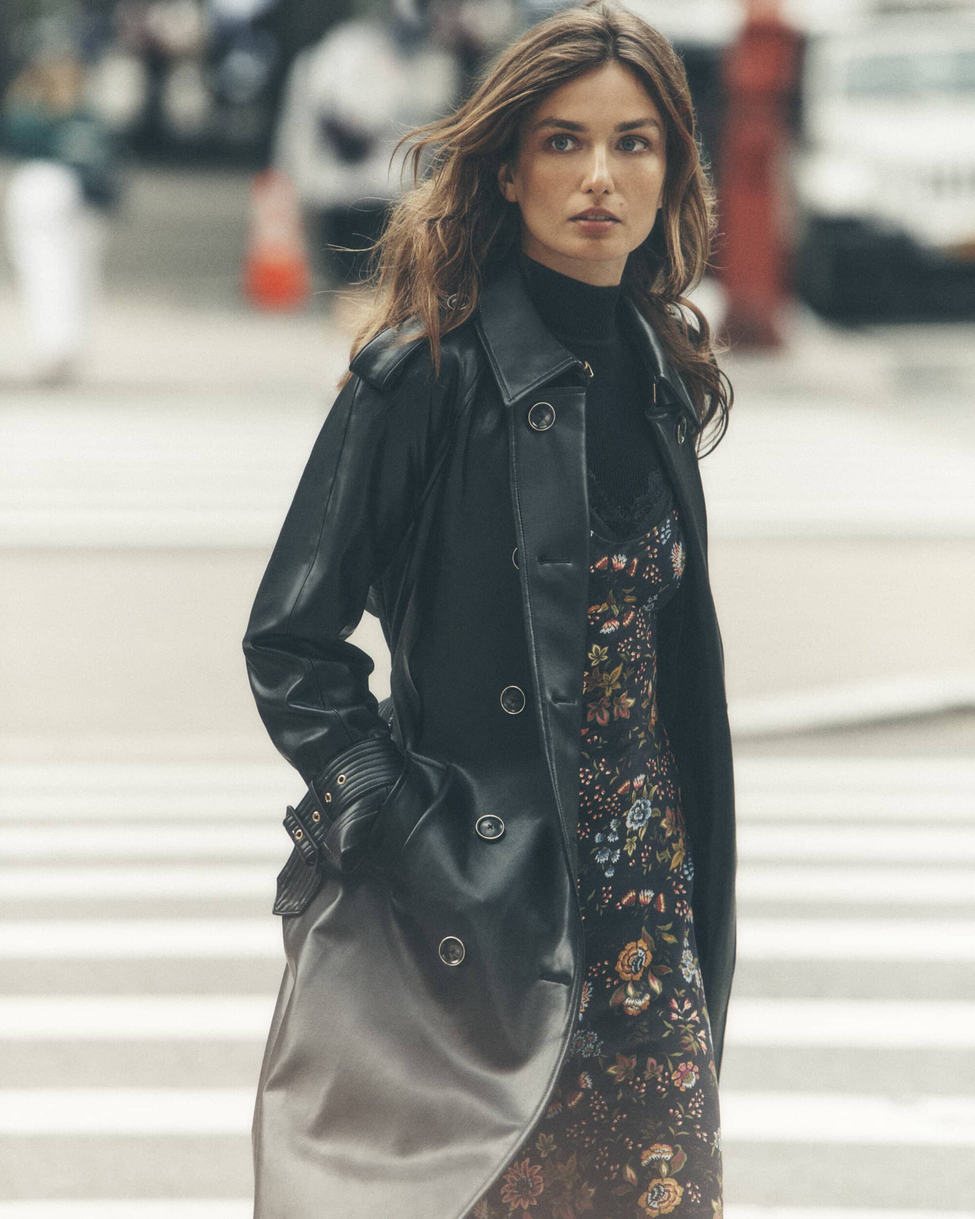 A woman in a black leather trench coat stands on a city street with crosswalk. She is wearing a floral dress and a black turtleneck.