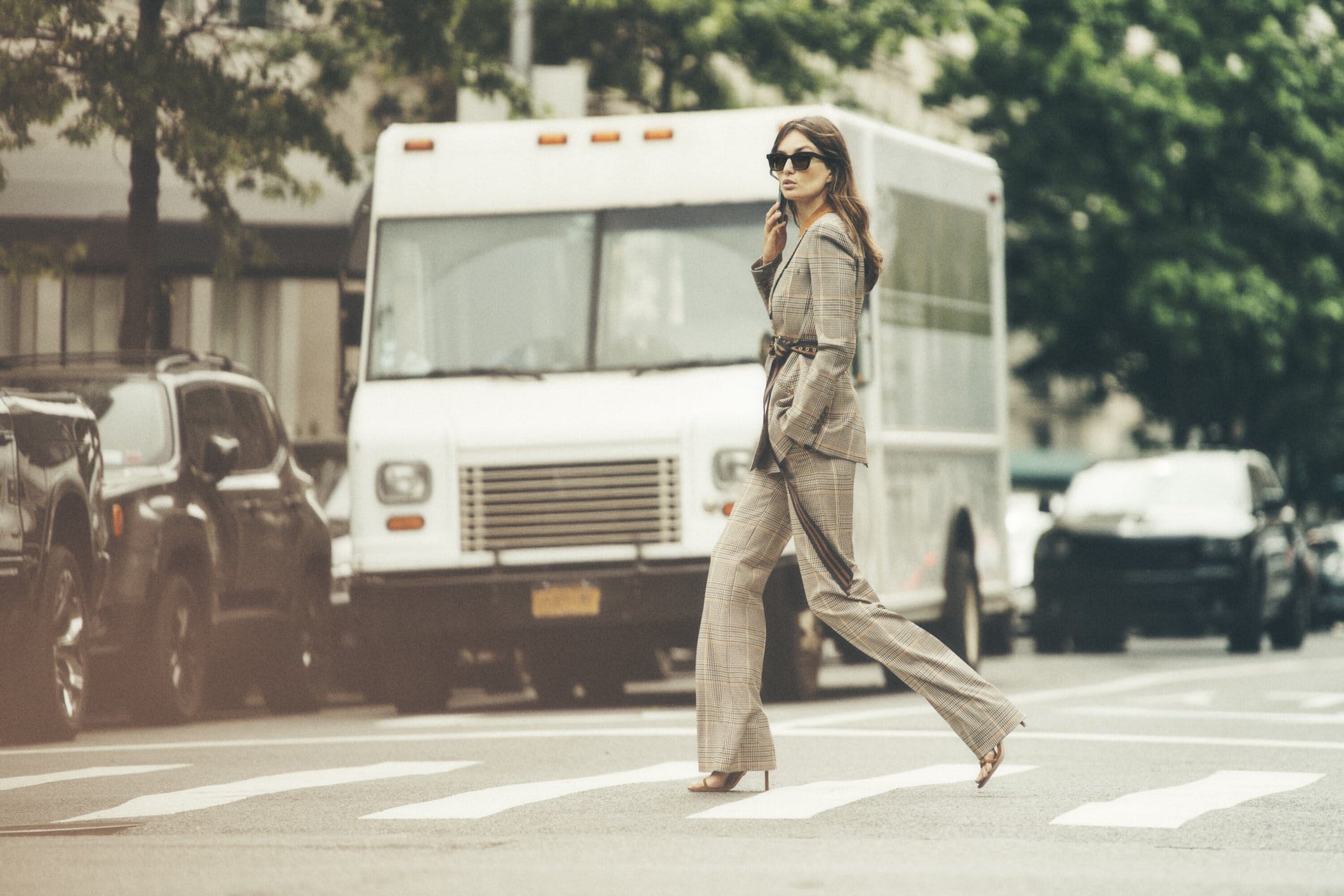 Woman in a plaid suit strides across a crosswalk on an urban street with a white van and other cars in the background.