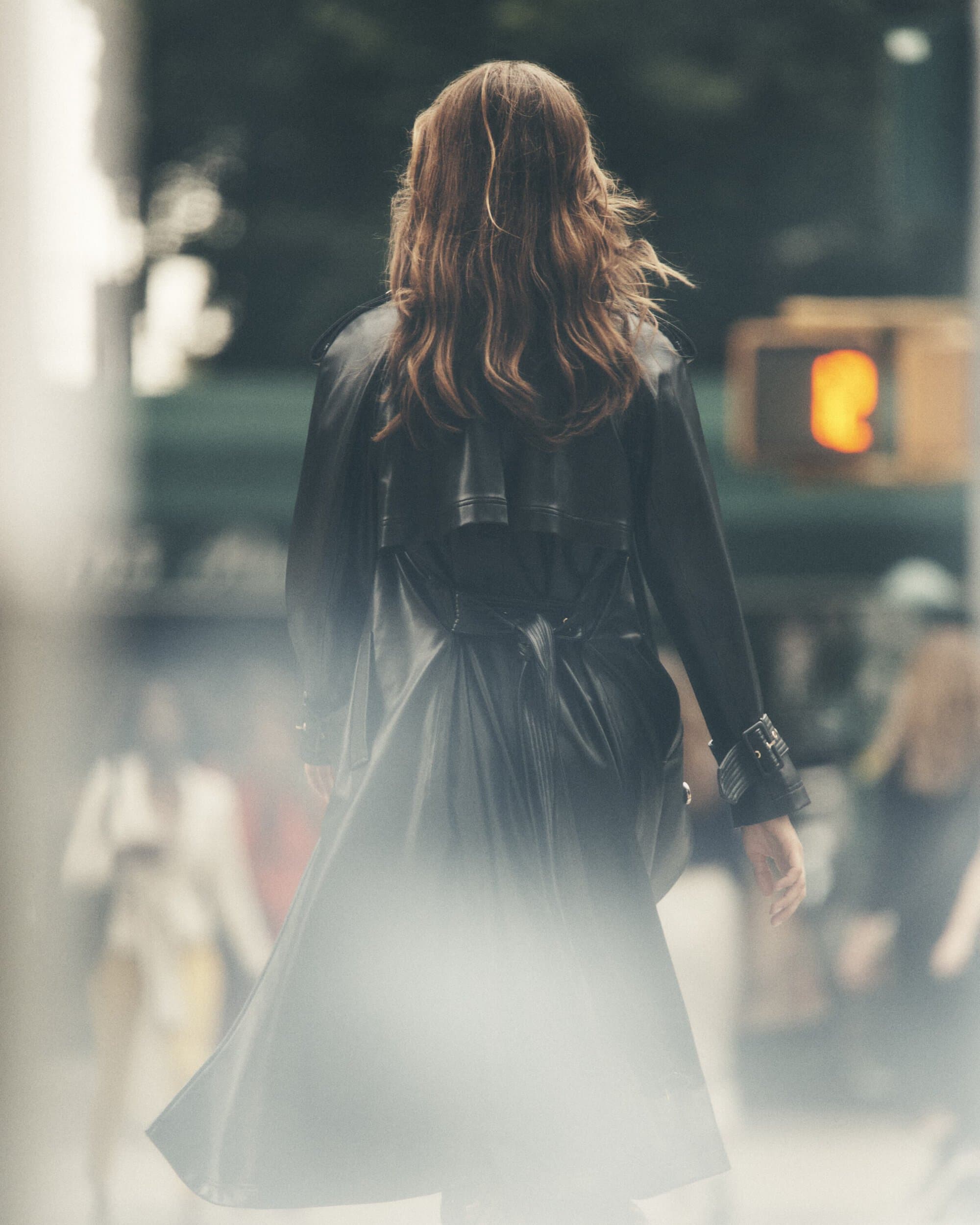Back view of a woman with long brown hair wearing a black trench coat walking on a city street with blurred pedestrians and buildings.