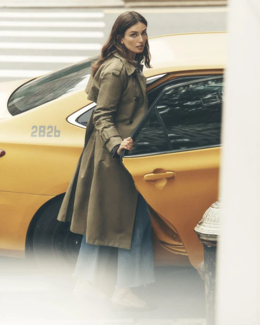 Woman in trench coat exiting yellow taxi on urban street. Fashion shot with natural light, medium framing, and shallow depth of field.