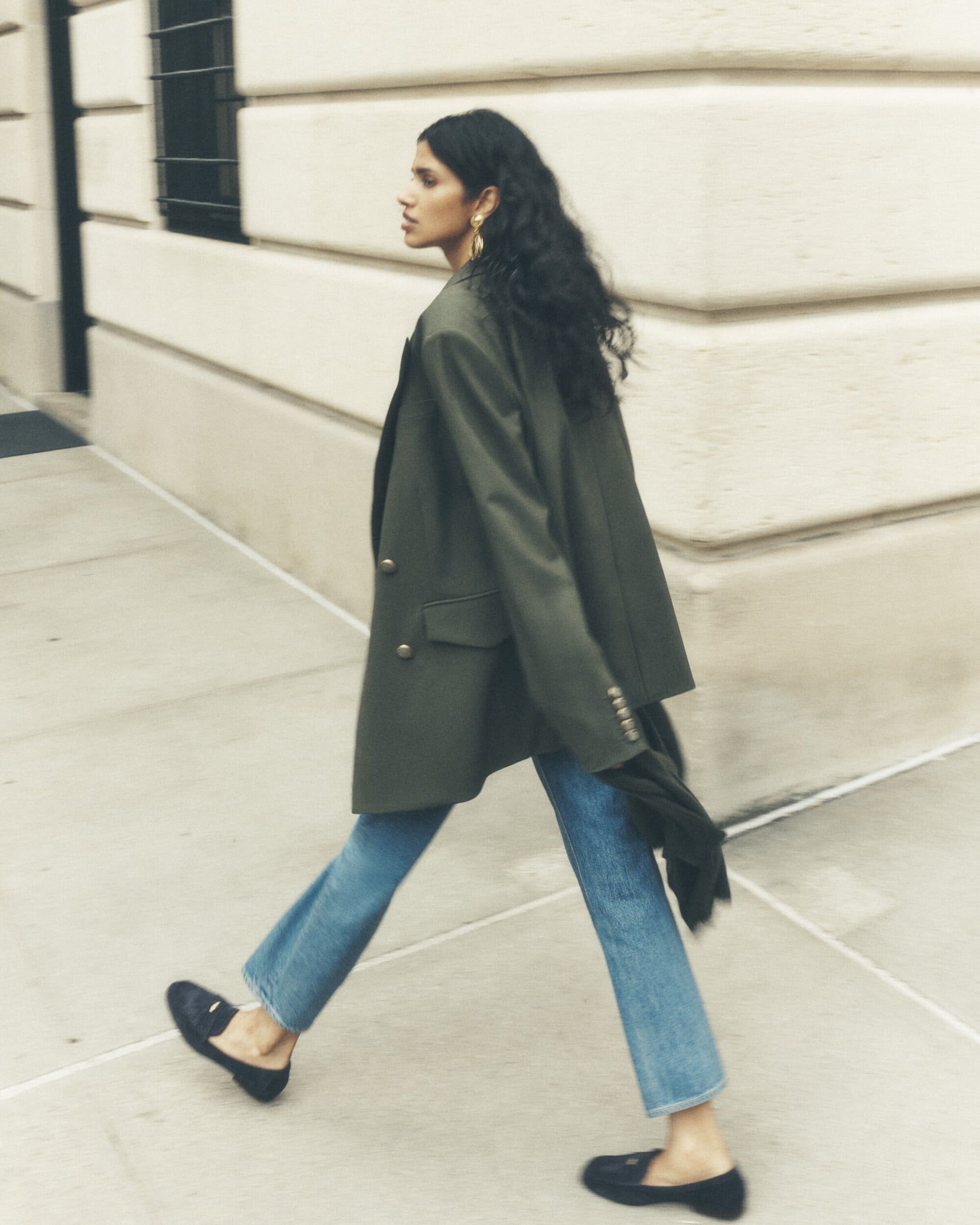 Woman in an olive blazer and jeans walks along a city street. Her black, wavy hair and a building in the background add visual interest.